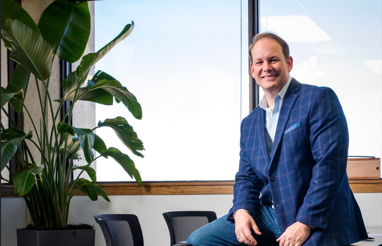 AmeriVet's CEO, Thomas Thill, sitting on top of a table, smiling. He is in a conference room with a potted plant in the background.